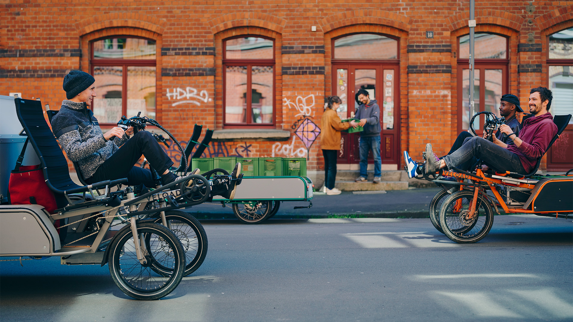 Zwei Lastenräder auf einer Straße, und Personen mit Gemüsekisten in Hintergrund